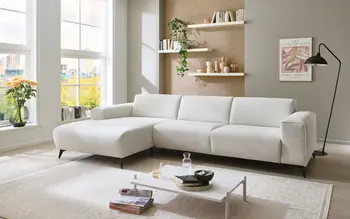 Stilvolles weißes Ecksofa in modernem Wohnzimmer mit großen Fenstern, Wandregalen und Dekoration. Rechts daneben lächelnde Frau, die ein Herz mit den Händen formt – Symbol für Wohlfühlen und Wohnkomfort.